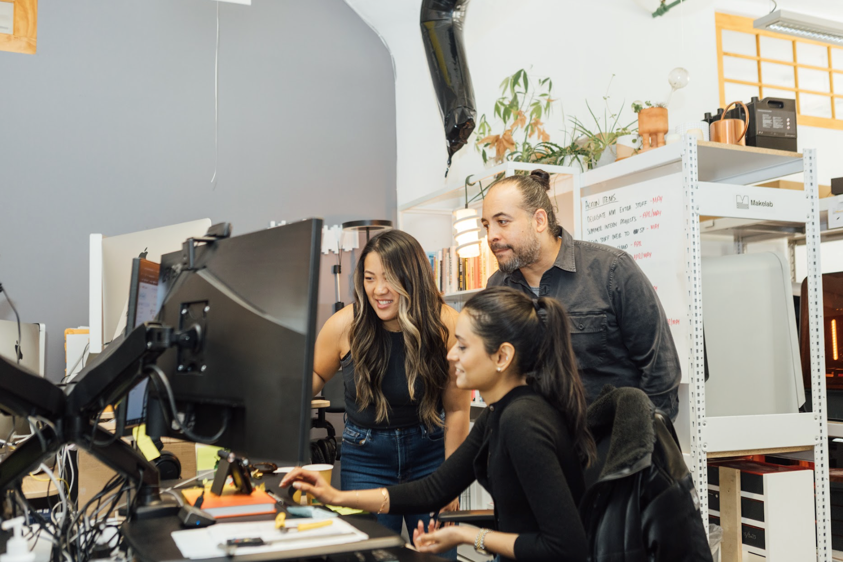 Three people looking at a large black computer monitor in an office, in front of a white bookshelf with the words “MAKELAB” printed on the side.